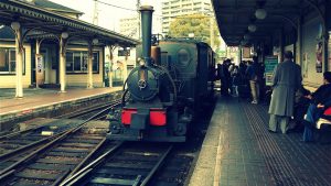 A vintage steam locomotive is stopped at a railway station platform, with several people waiting nearby. The nostalgic scene captures the charm of an old-fashioned Matsuyama tour, set in an urban area with buildings in the background.