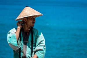 A man wearing a traditional straw hat and blue robe stands by the sea, looking to the side with one hand touching his neck—a serene scene inviting you to travel to Shikoku against a bright blue sea background.