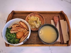 A wooden tray with a bowl of grilled pork over rice, chopped greens, and pickled plum, plus soup and pickled cabbage—perfect for savouring local flavours on your Matsuyama tour. Wooden chopsticks and spoon rest neatly on a serviette.