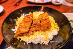 A black bowl filled with white rice, shredded egg, and grilled slices of glazed eel, served on a wooden table—perfect for enjoying during a Matsuyama tour with other local dishes in the background.