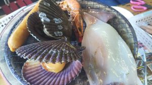 A plate with various fresh seafood, including scallops, a prawn, a squid, and a sausage, ready for cooking—perfect for those who travel to Shikoku and want to savour the region's authentic flavours. Some shells are open, revealing the meat inside.