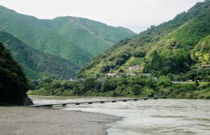 A narrow bridge spans a wide river surrounded by lush green hills, with a small village nestled among the dense trees in the background—inviting you to travel to Shikoku under a cloudy sky.