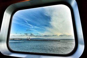 View of a calm sea, distant shoreline, and partly cloudy blue sky seen through a large, rectangular ship window with a metallic frame—an inviting scene for those who wish to travel to Shikoku.