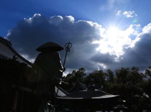 A silhouette of a statue holding a staff stands before traditional Japanese temple rooftops, with dramatic clouds and bright sunlight streaming through the sky—capturing the spirit of travel to Shikoku.