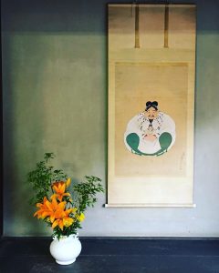 A white vase with orange lilies and green foliage sits on the floor next to a hanging scroll featuring a seated figure in traditional attire—capturing the tranquil essence of travel to Shikoku, all set against a plain grey wall.