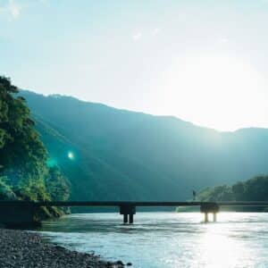 A person crosses a narrow bridge over a wide river, surrounded by lush green mountains in bright sunlight, with the sun low in the sky and a pebble shore in the foreground.